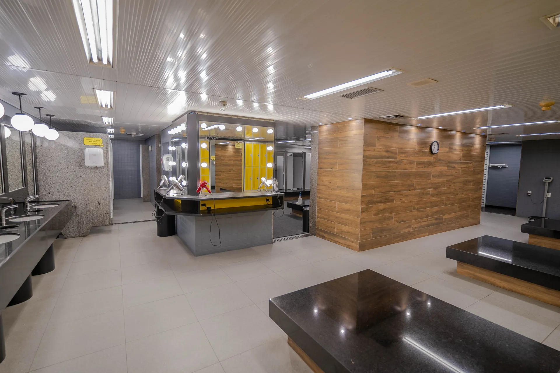 Vestiário da The Best, com bancos de granito, bancada de pias, espelhos iluminados e secadores