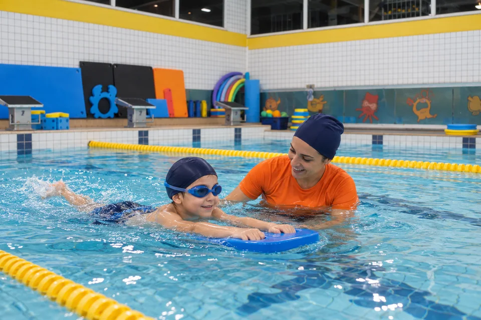 Criança praticando com prancha e orientação da professora na piscina infantil