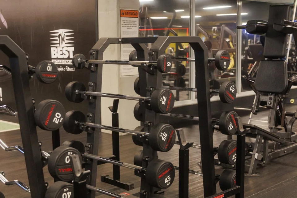 Rack de pesos livres com máquinas de musculação ao fundo na The Best